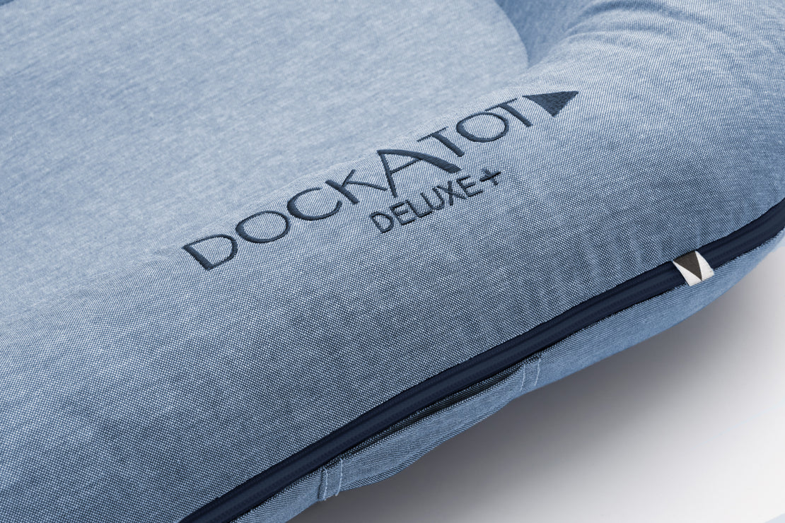 Close-up of a blue DockATot Deluxe+ Dock - Indigo Chambray multifunctional docking station showing the brand name embossed in gray on the fabric, with a focus on the textured material and the visible zipper detail along its edge.