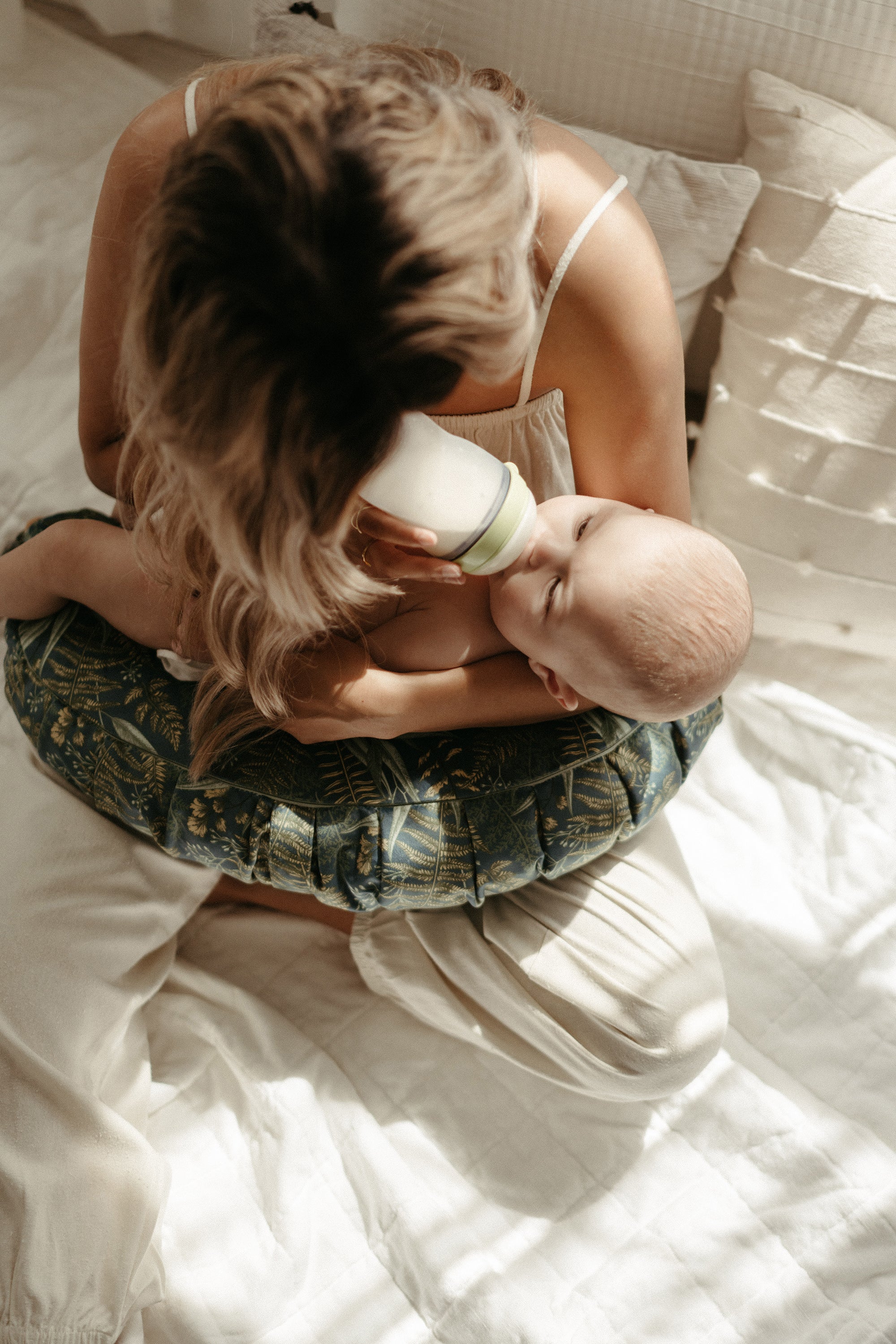 A mother sits on a bed feeding her baby with a bottle, using a DockATot nursing pillow for support. She&#39;s wearing a green patterned skirt and a white top, with sunlight filtering through creating a warm atmosphere.