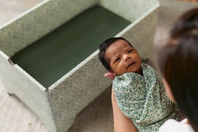 A newborn baby wrapped in a floral blanket looks towards the camera, held by a caregiver whose face is partially visible. They are seated near a DockATot Kind Essential Bassinet - Willow Boughs with ornately crafted pedestal feet in a softly.