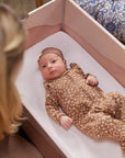 A newborn baby lies in a pink DockATot Kind Essential Bassinet with a white sheet, dressed in a brown onesie with a floral pattern, gazing upward. A woman, visible from the back and partially blurred.